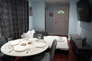 A hotel room with a dining table set for two, a bed with white linens, a television mounted on the wall, and a door with a window.