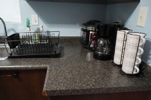 A kitchen counter with a coffee maker, a kettle, a dish rack, and a stack of mugs.