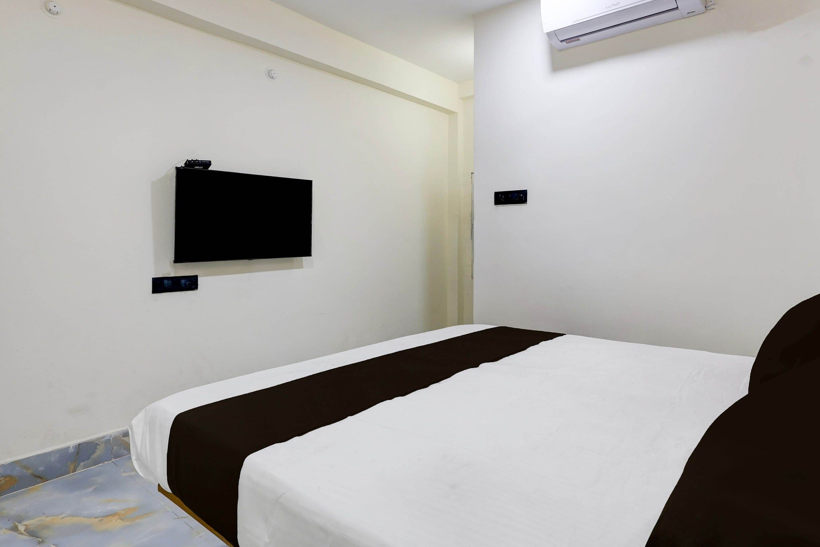 A hotel room with a single bed, a flat-screen TV mounted on the wall, and a small air conditioning unit above the bed.