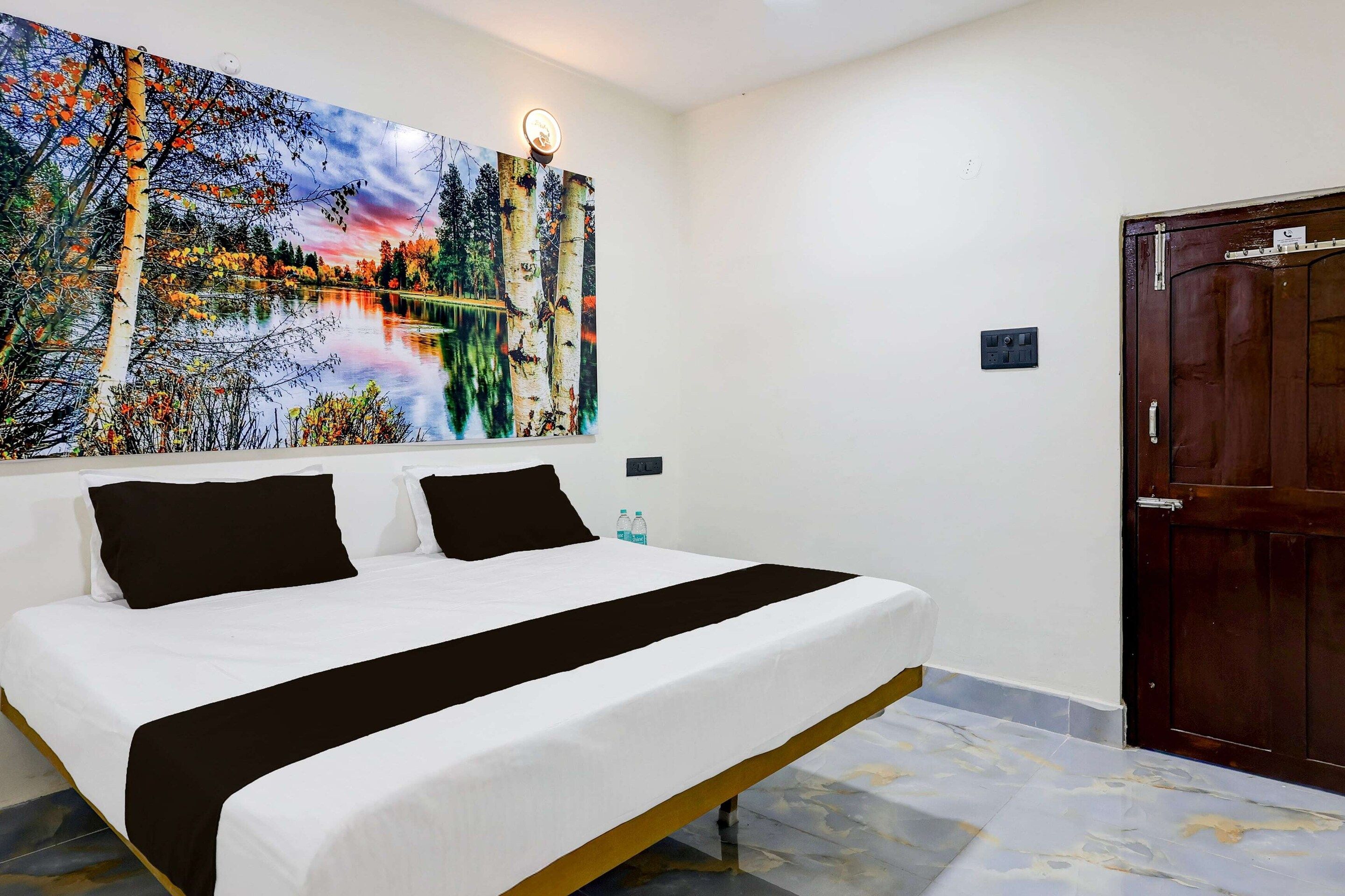 Quarto de hotel com cama, uma grande pintura de paisagem, uma porta e uma tomada.