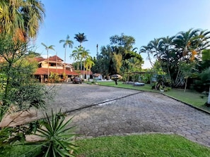 A paved courtyard with a building in the background, surrounded by palm trees and greenery. - Vila Palmar Hospedagem (Vassouras)