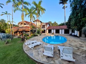 A pool area with lounge chairs, a thatched-roof structure, and a house with a red-tiled roof. - Vila Palmar Hospedagem (Vassouras)