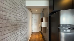 A narrow hallway with a brick wall on one side and a wooden floor. A modern stainless steel refrigerator is visible on the right. - Habitarte | 2 suites with views and leisure facilities high up (São Paulo)