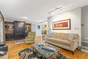 A living room with a sofa, armchair, and a glass coffee table. There is a bookshelf and a painting on the wall. - House on the Hill near Blue Ridge Pkwy & Downtown (Asheville)