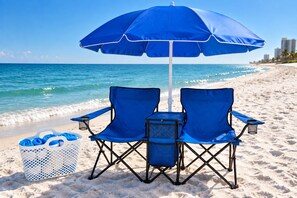 A beach scene with a blue umbrella, two blue folding chairs, and a white basket with blue towels.
