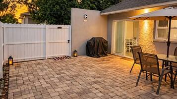 A backyard patio with a brick floor, a covered grill, a white fence, and a table with chairs.