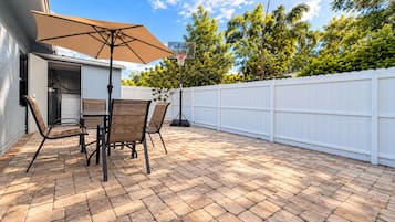 An outdoor patio with a brick floor, a table with chairs, and a white fence in the background.
