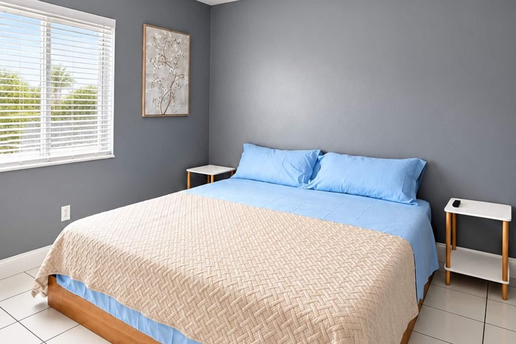 A bedroom with a bed, two bedside tables, a window with blinds, and a wall-mounted picture.