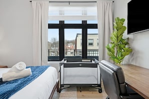 A hotel room with a bed, a desk with a chair, a large window with curtains, and a potted plant. - Penn 202| Queen Suite with Free parking + Sauna! (Pittsburgh)