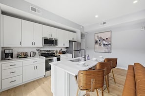 A modern kitchen with white cabinets, a microwave, stove, and a dining area with a brown leather couch and wooden chairs. - Penn 202| Queen Suite with Free parking + Sauna! (Pittsburgh)
