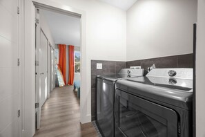 Een wasserette met twee wasmachines en een droger, een houten vloer, en een uitzicht door een geopende deur naar een balkon met oranje gordijnen.