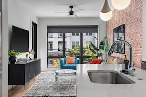A modern kitchen with a large window, a dining area with a blue sofa, and a visible balcony. - The Rhiannon | Pool, Parking & Private Balcony (Nashville)