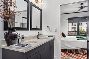 A modern bathroom with a large mirror, a vase with dried flowers, a hand soap dispenser, and a towel. - The Rhiannon | Pool, Parking & Private Balcony (Nashville)