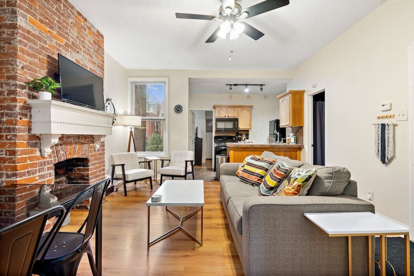 A living room with a brick fireplace, a ceiling fan, a sofa with colorful cushions, and a dining area with chairs and a table.