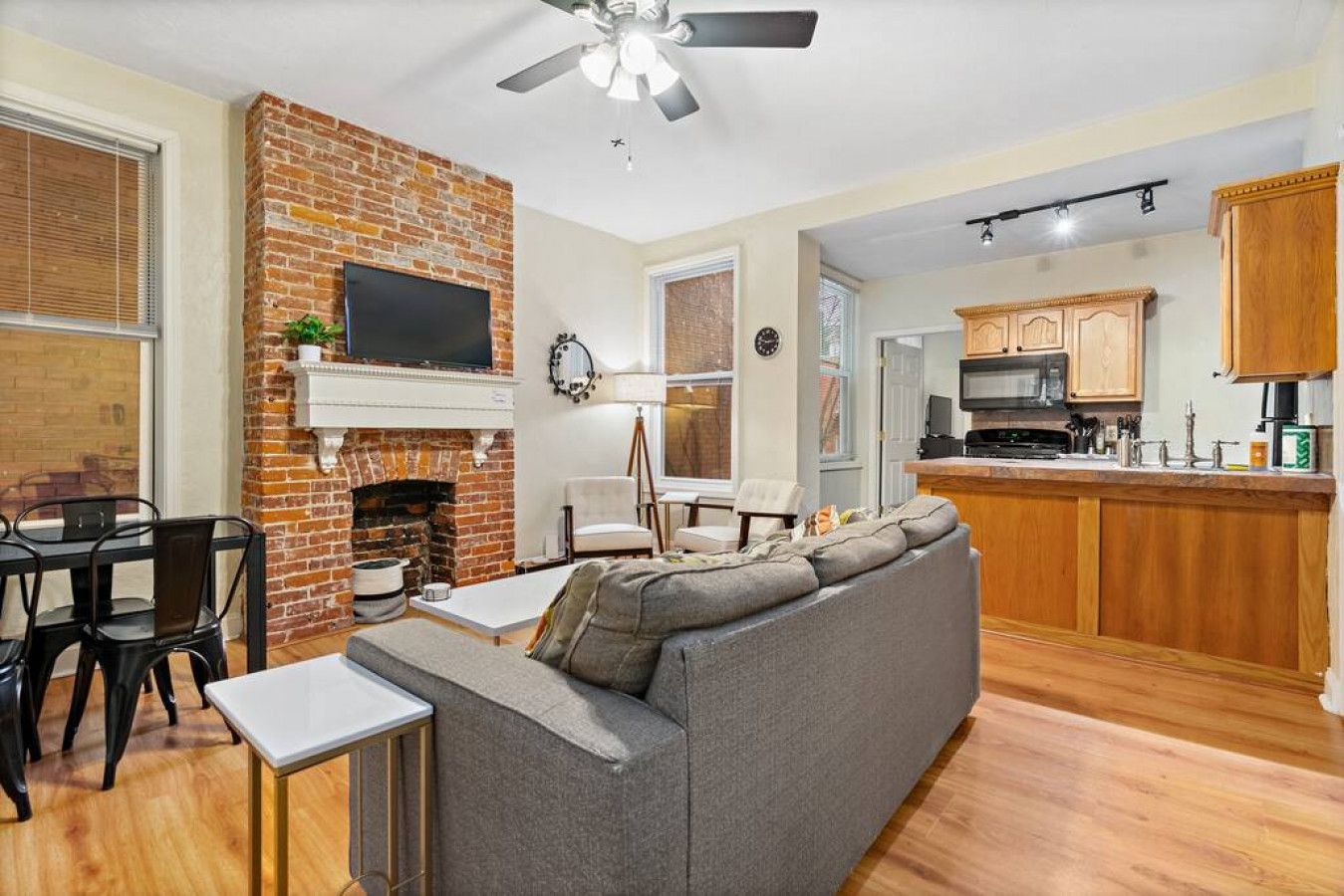 A living room with a gray sofa, a brick fireplace, a flat-screen TV, and a dining area with a table and chairs.