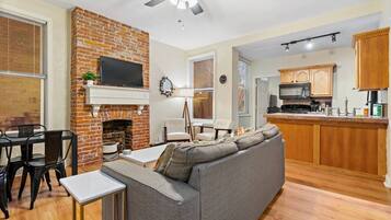 A living room with a gray sofa, a brick fireplace, a flat-screen TV, and a dining area with a table and chairs.