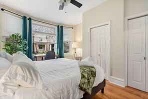 A hotel room with a bed, a desk, a chair, a lamp, and a view of the outside. - 5531 Kentucky APT 1 · Cozy 2BR ~ $0 cleaning fee & steps from Walnut St! (Pittsburgh)