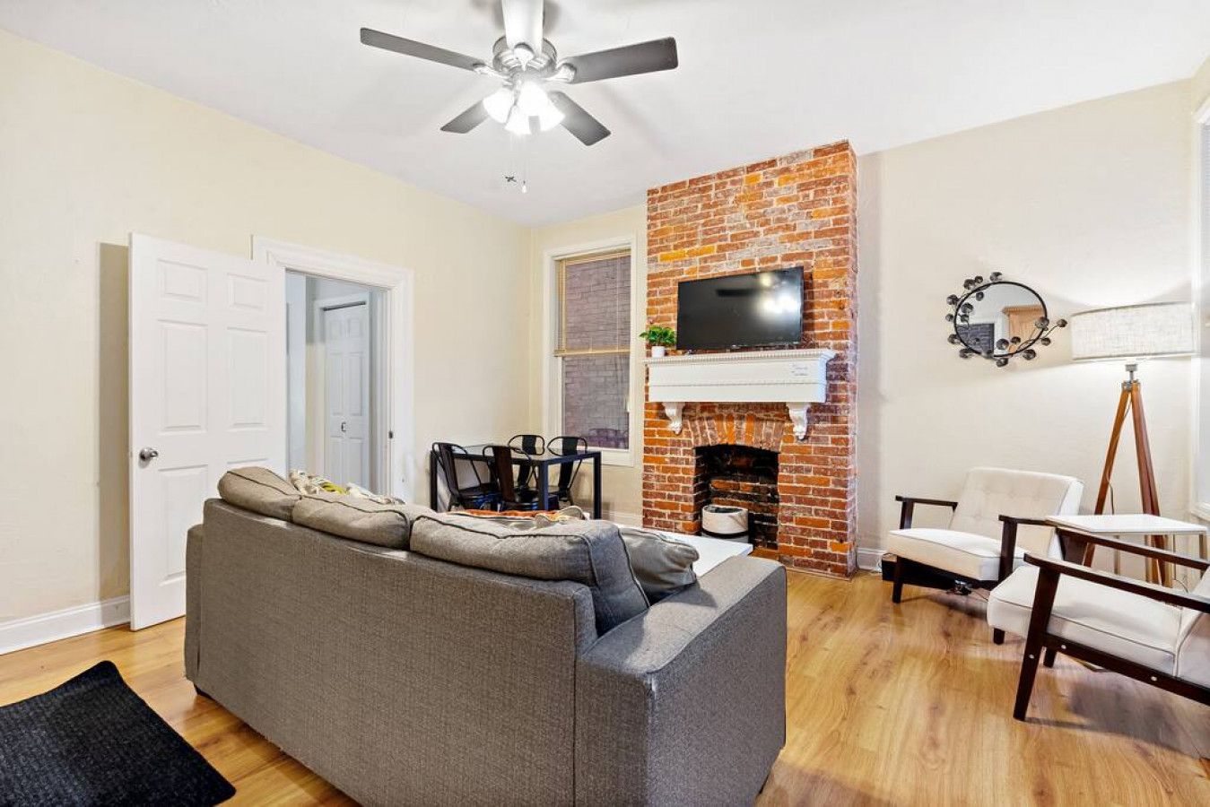 A living room with a gray sofa, a brick fireplace, a ceiling fan, and a TV mounted above the fireplace.