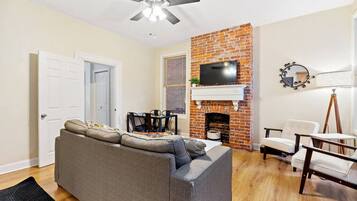 A living room with a gray sofa, a brick fireplace, a ceiling fan, and a TV mounted above the fireplace.