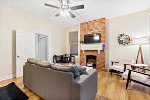 A living room with a gray sofa, a brick fireplace, a ceiling fan, and a TV mounted above the fireplace. - 5531 Kentucky APT 1 · Cozy 2BR ~ $0 cleaning fee & steps from Walnut St! (Pittsburgh)