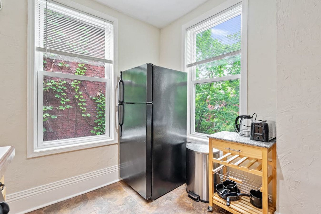 Cocina compacta con refrigerador de acero inoxidable, repisa de madera con utensilios de cocina y ventana con vista a follaje.