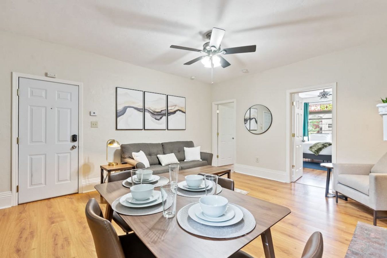 A dining area with a wooden table set for four, a grey sofa, and a ceiling fan.