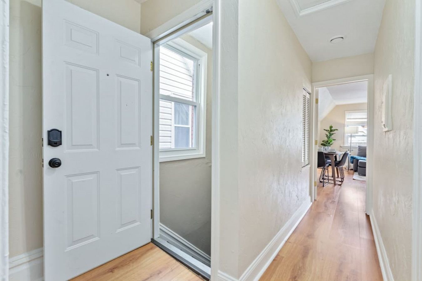 A hallway with a white door, a window, and a view into a dining area with a table and chairs.