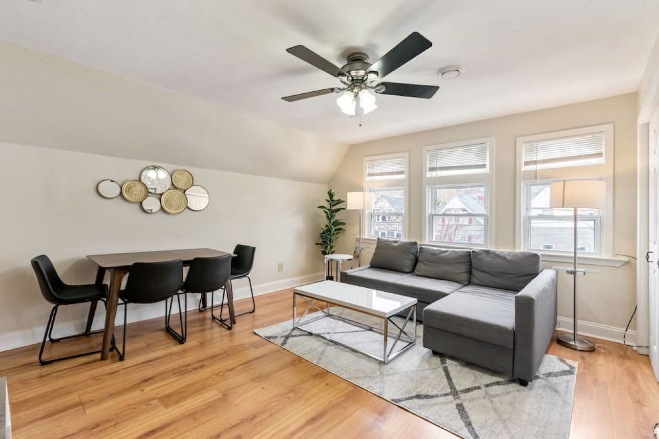 A living room with a dining table, chairs, a sofa, a coffee table, a ceiling fan, and a view of the outside through large windows.