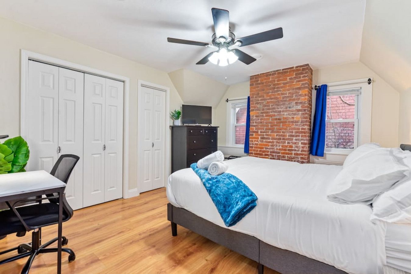 A bedroom with a bed, a desk, a chair, a ceiling fan, and a brick wall.