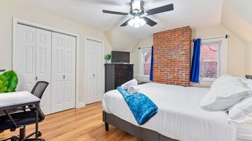 A bedroom with a bed, a desk, a chair, a ceiling fan, and a brick wall.