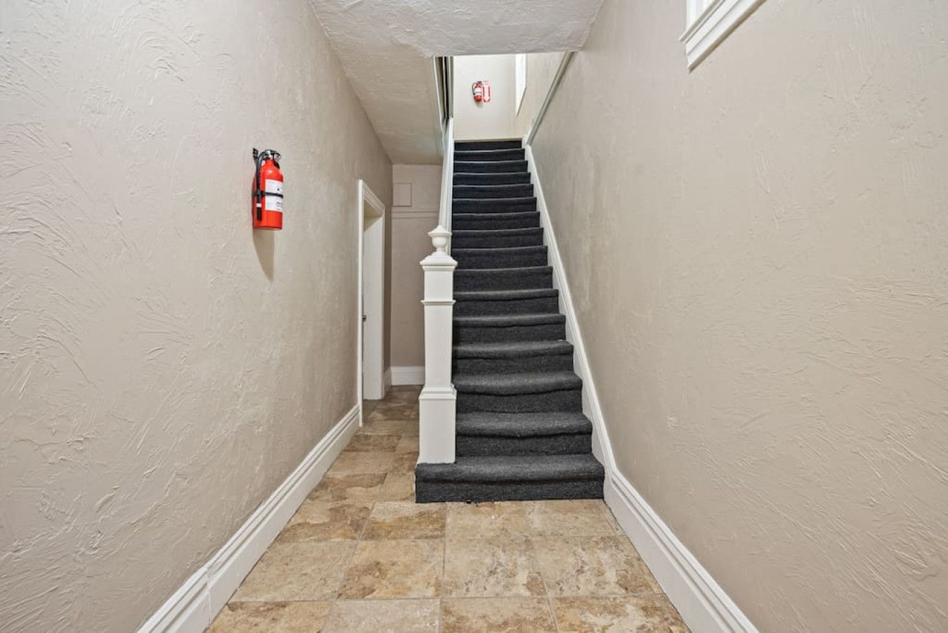 A hallway with a carpeted staircase, a fire extinguisher mounted on the wall, and a door at the end.
