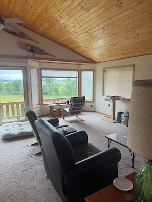 A cozy room with a wooden ceiling, a black leather sofa, a striped chair, a small table, and a view of a green landscape through large windows. - Star Lake Escape, Dent MN (Dent)
