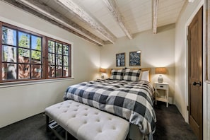 Ein Schlafzimmer mit einem großen Fenster, einem Bett mit karierter Bettdecke, einem Nachttisch mit Lampe und einer Tür.