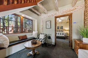 Ein gemütliches Zimmer mit einer Bank an einem großen Fenster, einem kleinen runden Tisch und einem Bett mit kariertem Überwurf.