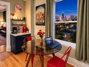 A modern dining area with a glass table, red chairs, a laptop, and a cityscape view through the window.