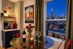 A dining area with a table set for two, a bottle of wine, and a candle. A cityscape view is visible through a window.