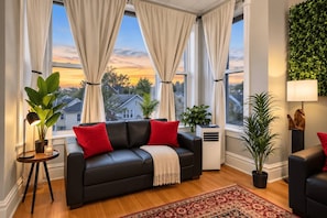 A living room with a black leather sofa, red cushions, a beige throw, a round table with a lamp, and a potted plant.
