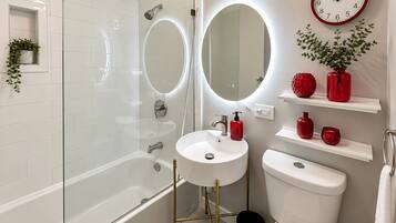 A modern bathroom with a glass-enclosed shower, a white sink, a toilet, and a round mirror with a red frame.