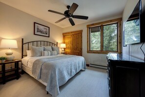 Ein Schlafzimmer mit einem Bett, zwei Nachttischlampen, einem Deckenventilator, einem Fenster mit Blick auf Bäume und einer Kommode.