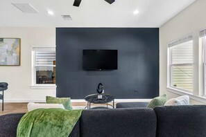 Ein modernes Wohnzimmer mit dunkler Akzentwand, einem wandmontierten Flachbildfernseher, einem runden Couchtisch und einem Sofa mit grüner Tagesdecke.