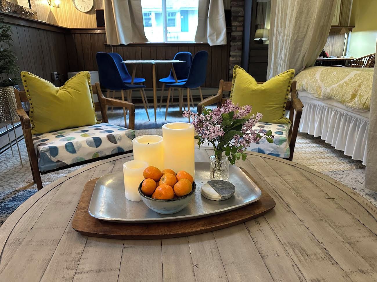 A cozy room with a round wooden table, a tray with candles, a bowl of oranges, and a vase with flowers.