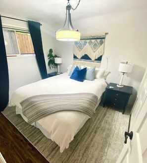 Un dormitorio con cama, mesitas de noche, lámpara colgante, ventana con cortinas y un tapiz en la pared.