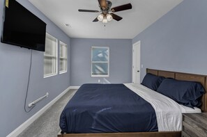 A bedroom with a bed, a ceiling fan, a TV mounted on the wall, and windows with blinds.