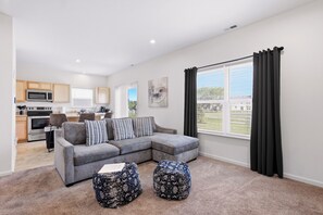 A modern living room with a grey sectional sofa, two patterned ottomans, a dining area with a table and chairs, and a kitchen with appliances. - The Aero   NEW!  Wifi, Parking, Massage Chair, Washer/Dryer (Indianapolis)
