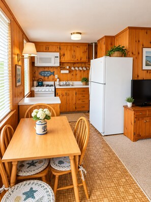 A compact kitchen with wooden cabinets, a white refrigerator, a stove, and a microwave. There is a dining table with chairs and a vase of flowers. - Oceanside Pearl-Cozy 2 Bdrm Coastal Getaway, 2/10th of a Mile from Beach (Dennis)