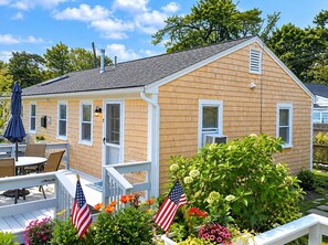 Ein Bungalow mit Veranda, kleiner Terrasse und Garten mit Blumen und zwei amerikanischen Flaggen.