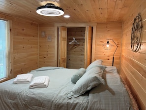 A wooden cabin room with a bed, a hanging light fixture, and a window with blinds. - The Hidden Bungalow sleeps 4, has  AC & internet on Wabasis Lake in GreenvilleMI (Greenville)