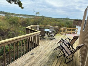Une terrasse en bois, aménagée avec du mobilier d’extérieur comprenant une table et des chaises, qui offre une vue sur un paysage naturel avec des arbres et une étendue d’eau.