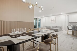 Un espace de repas moderne avec une longue table dressée pour six personnes, une cuisine en arrière-plan et un coin salon avec un canapé et une table basse.