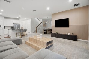 Un salon moderne avec un grand téléviseur à écran plat fixé au mur, un canapé, une table basse et un coin cuisine à l’arrière-plan.